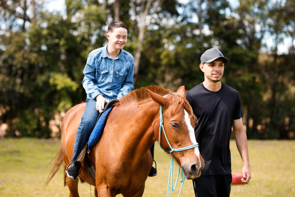 A young man with Down syndrome is happily riding a reddish-brown horse in a sunny, grassy field.