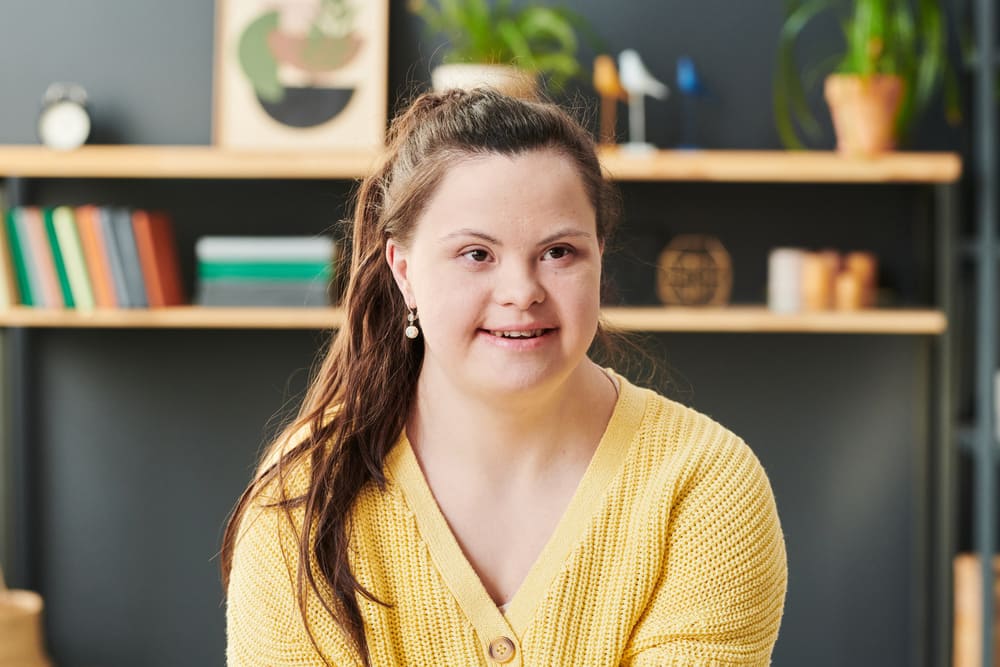 A young woman with Down syndrome, wearing a bright yellow cardigan, is looking directly at the camera with a friendly, gentle smile.