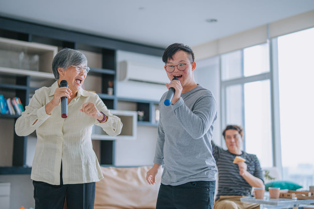 A woman with short grey hair and glasses, wearing a light yellow blouse and dark trousers, is enthusiastically singing into a microphone on the left.