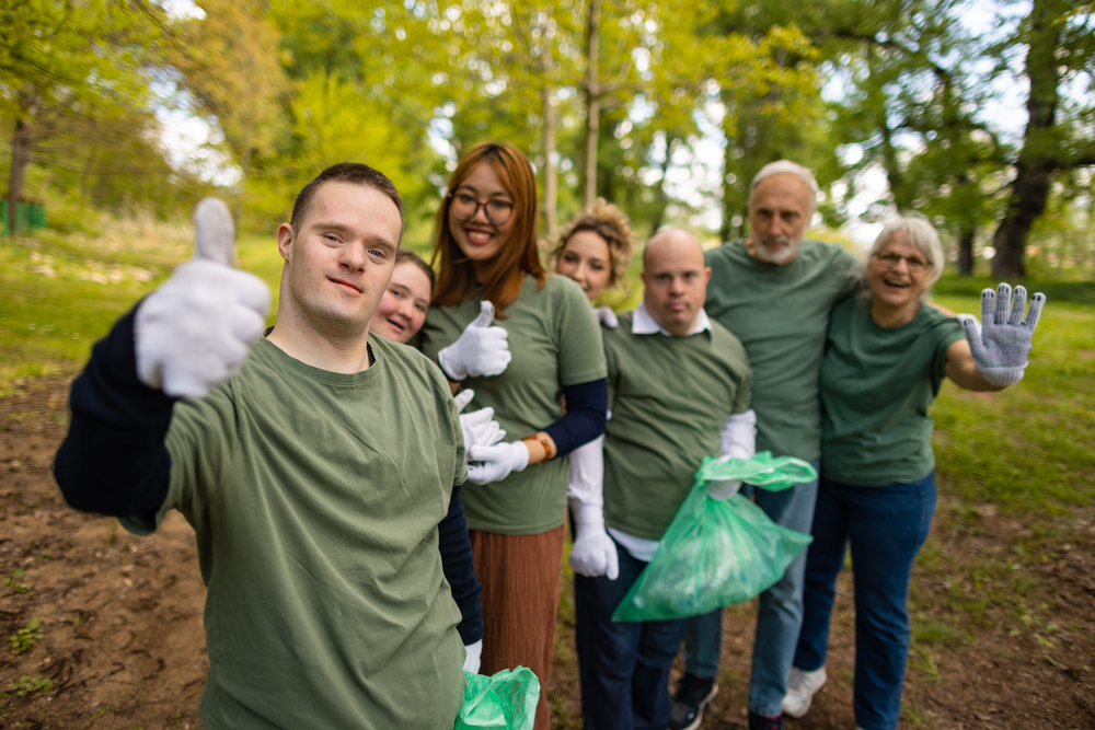 A group of different ages and backgrounds, all wearing green tops, are standing together outdoors in a slightly out-of-focus park or wooded area.